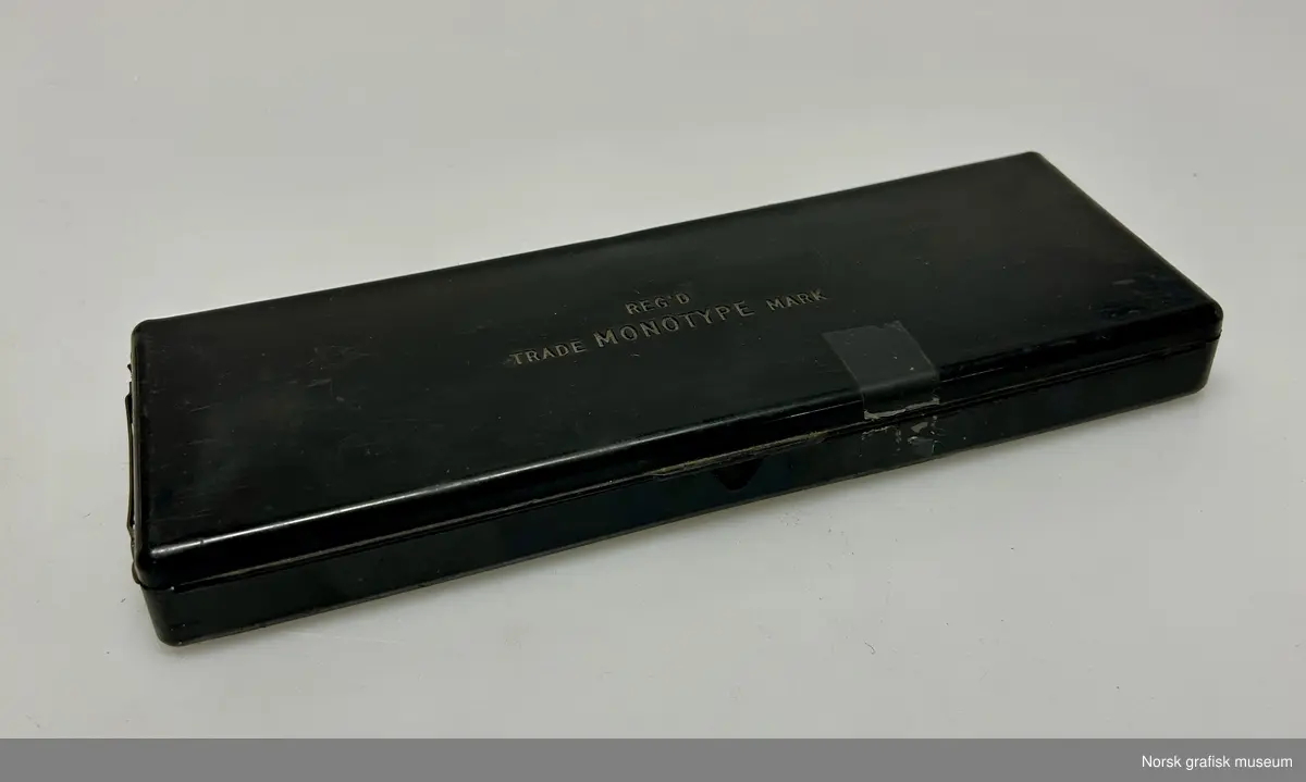 Et plastskrin inneholdende matriser til Monotype. De individuelle matrisene er plassert i små rom i skrinet. På undersiden av lokket et det limt et skjema. Firmanavet er preget inn i lokket.