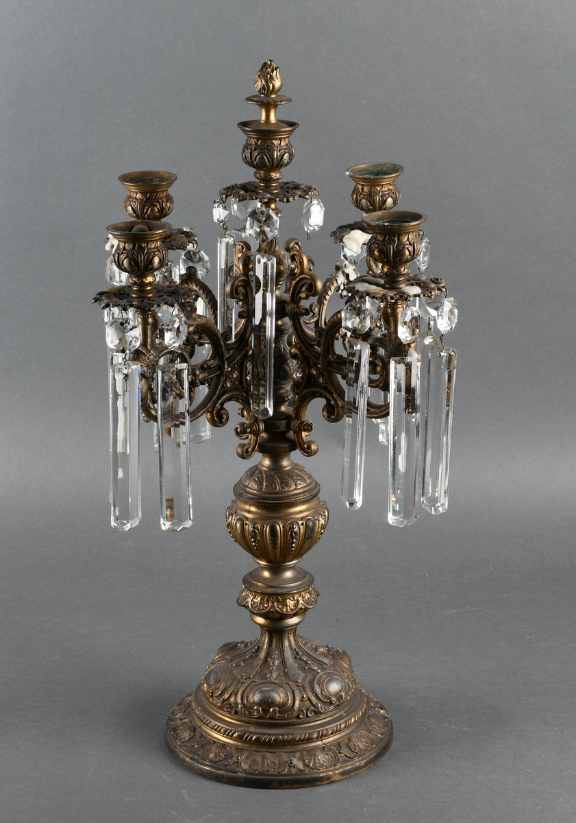 Kandelaber av messing med prismer hengende på. Fire lysholdere med prismer rundt mansjettene. I midten går selve staken opp i en lysholder med avtagbar propp. Foten er sterkt profilert i rikt mønstret støpegods. Det mangler noen prismer.