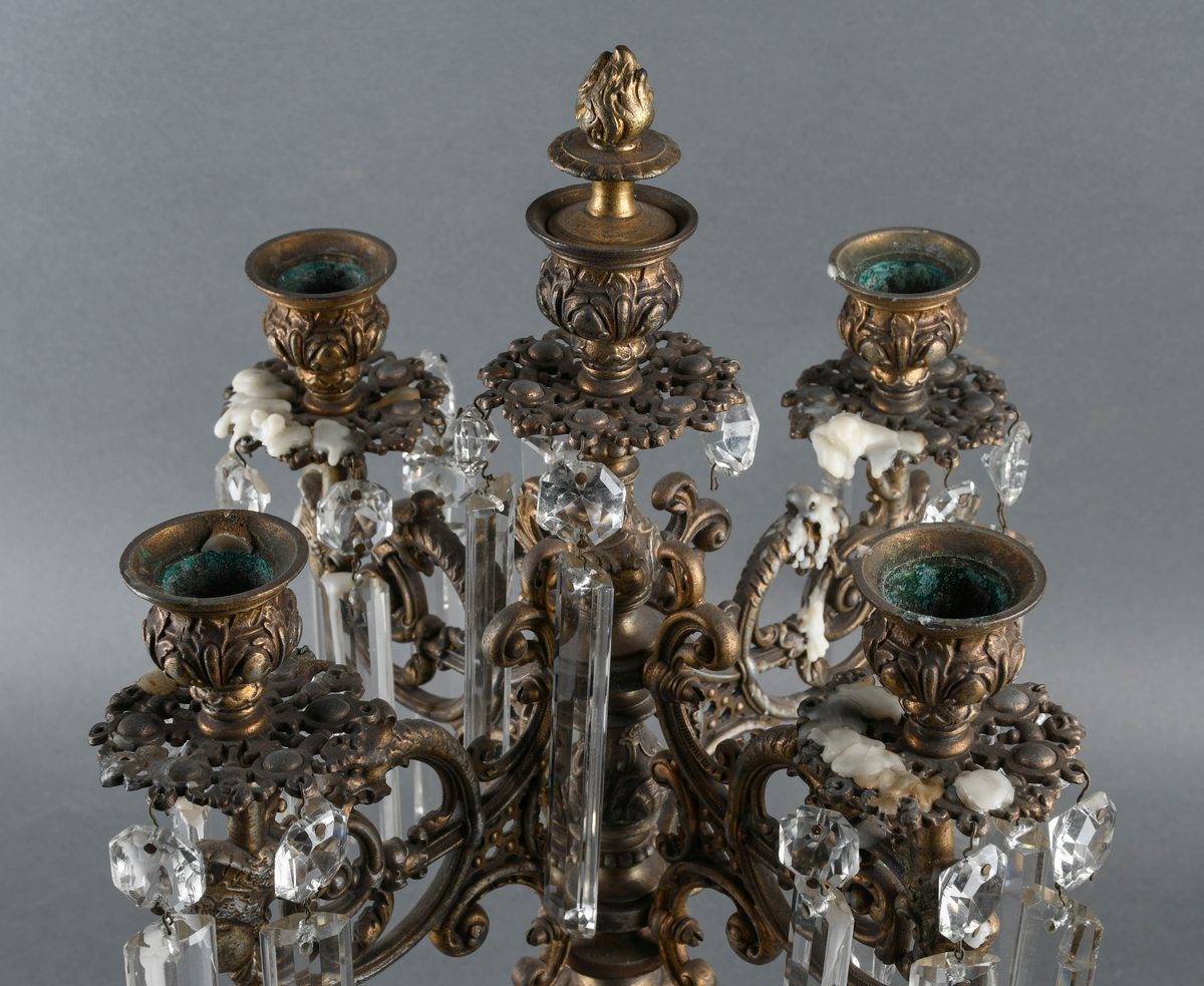 Kandelaber av messing med prismer hengende på. Fire lysholdere med prismer rundt mansjettene. I midten går selve staken opp i en lysholder med avtagbar propp. Foten er sterkt profilert i rikt mønstret støpegods. Det mangler noen prismer.