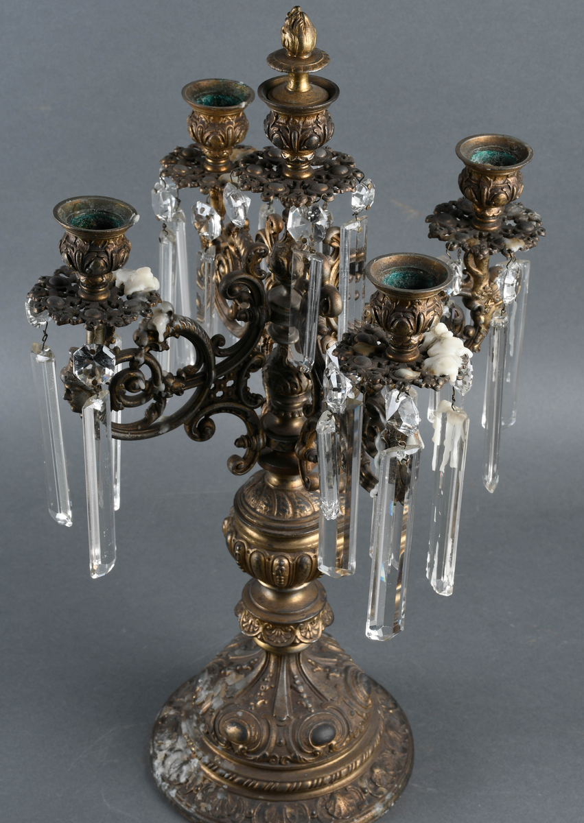 Kandelaber av messing med prismer hengende på. Fire lysholdere med prismer rundt mansjettene. I midten går selve staken opp i en lysholder med avtagbar propp. Foten er sterkt profilert i rikt mønstret støpegods. Det mangler noen prismer.