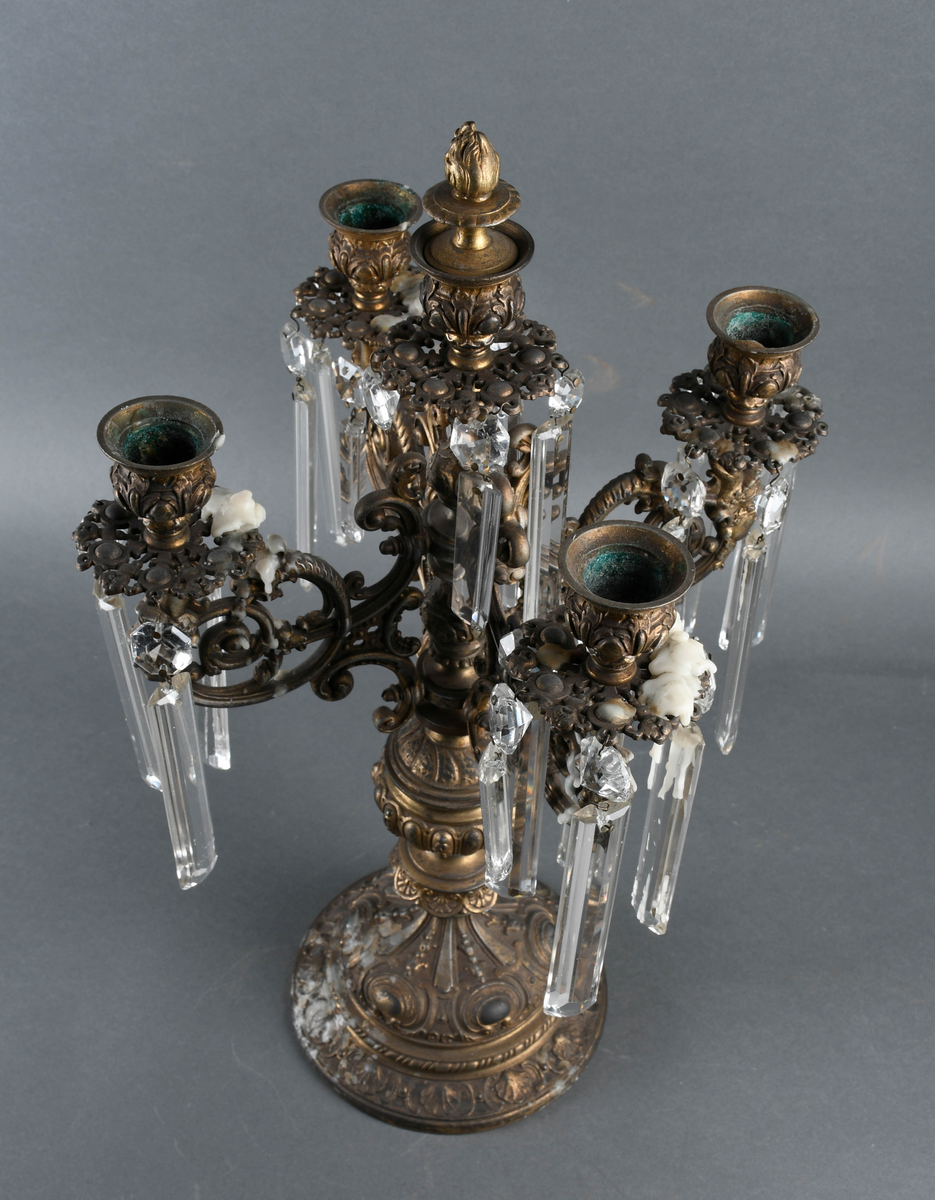 Kandelaber av messing med prismer hengende på. Fire lysholdere med prismer rundt mansjettene. I midten går selve staken opp i en lysholder med avtagbar propp. Foten er sterkt profilert i rikt mønstret støpegods. Det mangler noen prismer.