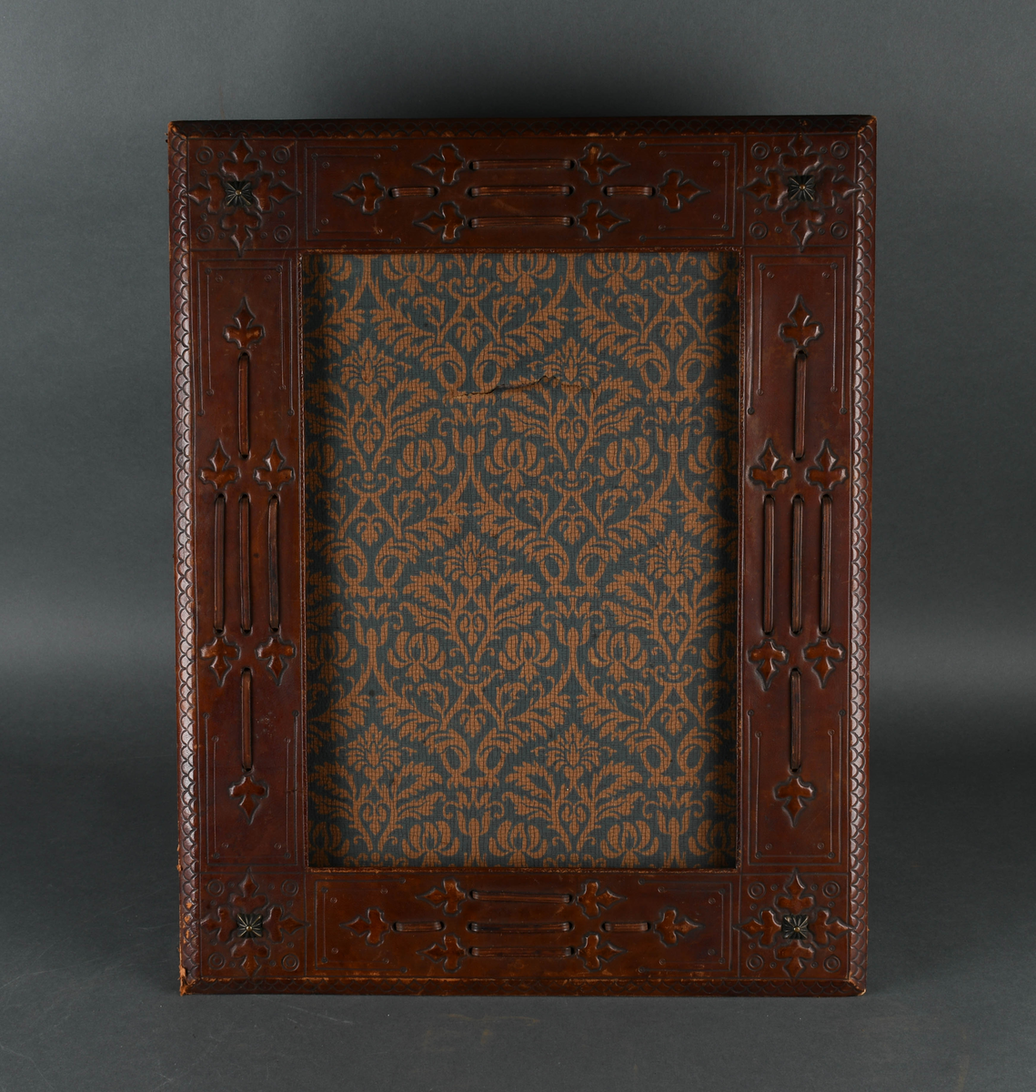 Rektangulær bilderamme av tre trukket med lær. Læret har punslet og hevet dekor i form av treflikete blader og geometriske mønstre, samt reimer. I hvert hjørne er en firkantet, rillet metallstift. Bakdelen er hengslet øverst med en støttende fot. Innsiden er kledd med papir med symmetrisk plantedekor.