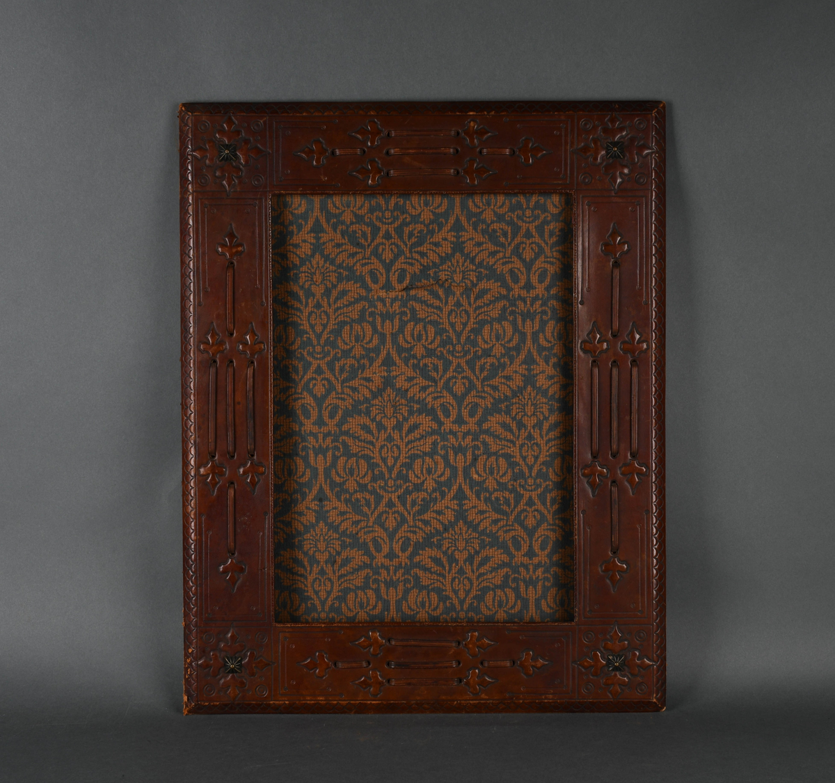 Rektangulær bilderamme av tre trukket med lær. Læret har punslet og hevet dekor i form av treflikete blader og geometriske mønstre, samt reimer. I hvert hjørne er en firkantet, rillet metallstift. Bakdelen er hengslet øverst med en støttende fot. Innsiden er kledd med papir med symmetrisk plantedekor.