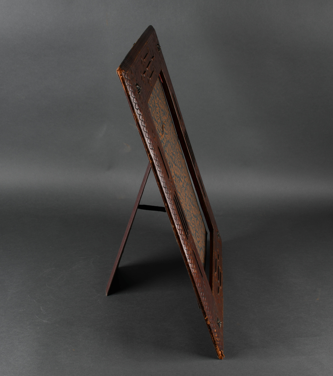 Rektangulær bilderamme av tre trukket med lær. Læret har punslet og hevet dekor i form av treflikete blader og geometriske mønstre, samt reimer. I hvert hjørne er en firkantet, rillet metallstift. Bakdelen er hengslet øverst med en støttende fot. Innsiden er kledd med papir med symmetrisk plantedekor.