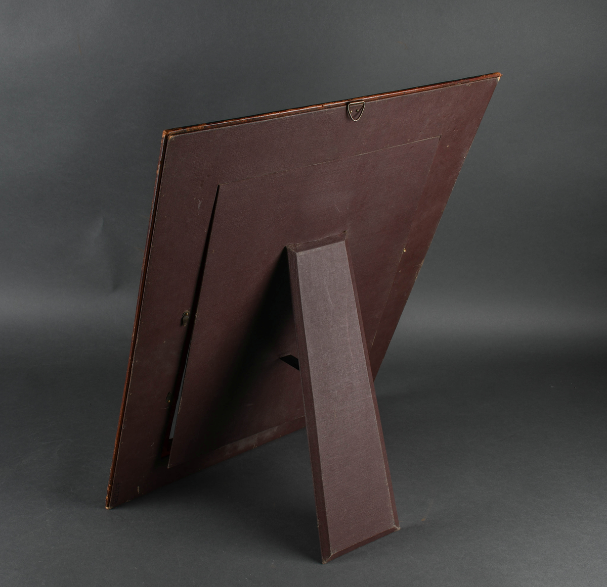 Rektangulær bilderamme av tre trukket med lær. Læret har punslet og hevet dekor i form av treflikete blader og geometriske mønstre, samt reimer. I hvert hjørne er en firkantet, rillet metallstift. Bakdelen er hengslet øverst med en støttende fot. Innsiden er kledd med papir med symmetrisk plantedekor.