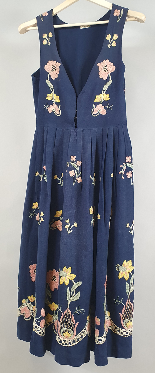 Blå bunad i tynt ullstoff, og med brodering på liv og stakk. Mønster av store gule og rosa blomster langs skjørtekant, på ryggen og forstykkene, og små blomster spredd utover skjørt og liv. 
Åpning i venstre side under ermet. Metallhemper i åpning foran. Livet er fôret med et blått syntetisk stoff.