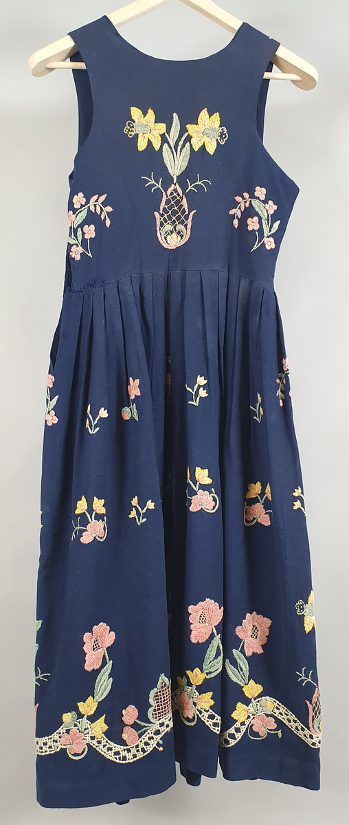 Blå bunad i tynt ullstoff, og med brodering på liv og stakk. Mønster av store gule og rosa blomster langs skjørtekant, på ryggen og forstykkene, og små blomster spredd utover skjørt og liv. 
Åpning i venstre side under ermet. Metallhemper i åpning foran. Livet er fôret med et blått syntetisk stoff.