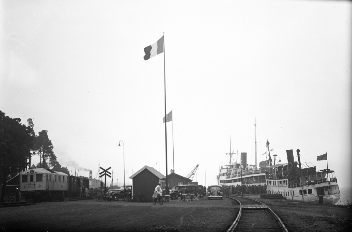 Hamnen, Gotlandskajen. Till vänster ett lok med tågvagnar, till höger syns två fartyg liggande vid kaj. Det ena är gotlandsbåten M/S Gotland och det närmaste är Eskilstuna Express. På kajen står bilar parkerade, människor ombord på Gotlandsbåten som ligger längre bort. Flaggor är hissade, den närmaste flaggan är inte svensk.
