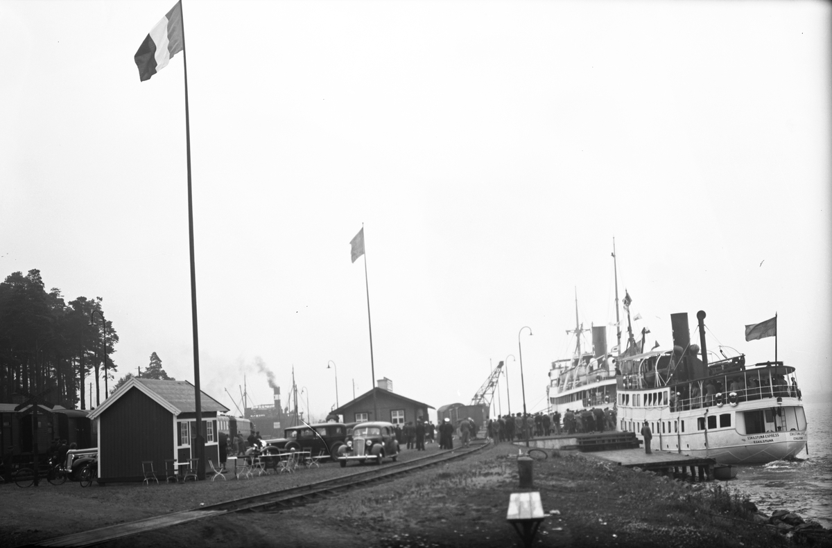 Hamnen, Gotlandskajen. Två fartyg ligger vid kaj, det ena är M/S Gotland och det andra Eskilstuna Express. På kajen står bilar parkerade, människor ombord på Gotlandsbåten som ligger längre bort. Flaggor är hissade, den närmaste flaggan är inte svensk.