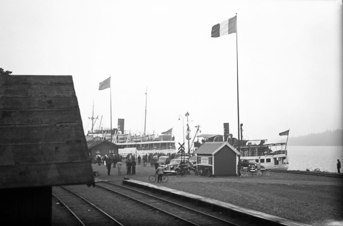 Hamnen, Gotlandskajen. Till vänster ett lok med tågvagnar, till höger syns två fartyg liggande vid kaj. Det ena är gotlandsbåten M/S Gotland och det närmaste är Eskilstuna Express. På kajen står bilar parkerade, människor ombord på Gotlandsbåten som ligger längre bort. Flaggor är hissade, den närmaste flaggan är inte svensk.