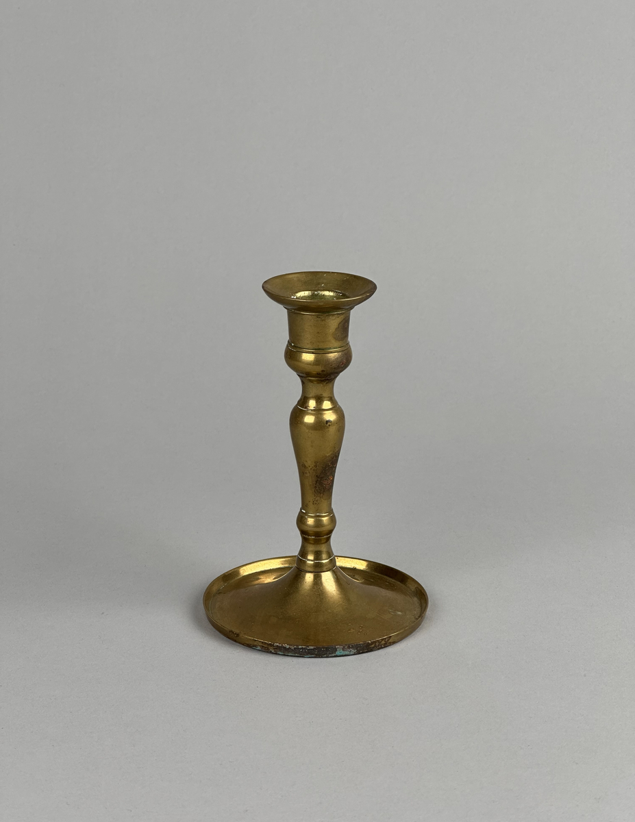 Ljusstake av mässing, i två delar. Lite skålformat brätte på ljuspipan. Svagt balusterformad stake med gängad fot. Fotens kant är tvärt uppvikt.