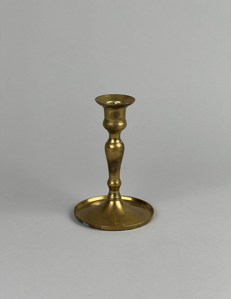 Ljusstake av mässing, i två delar. Lite skålformat brätte på ljuspipan. Svagt balusterformad stake med gängad fot. Fotens kant är tvärt uppvikt.