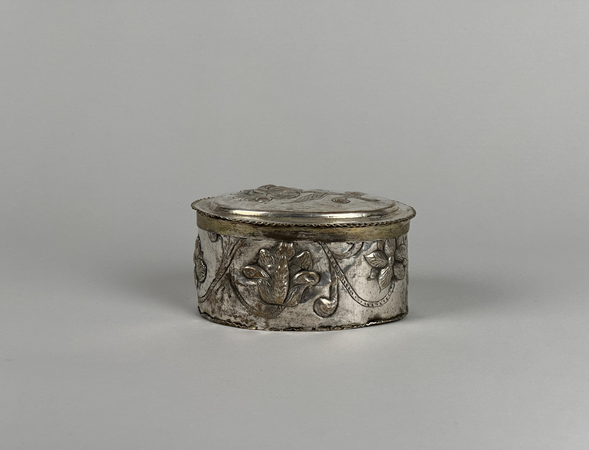 Oblatask tillverkad i försilvrad koppar, oval form med driven ornering. Lock dekorerad med en ros och sidorna med en girlang av rosor.