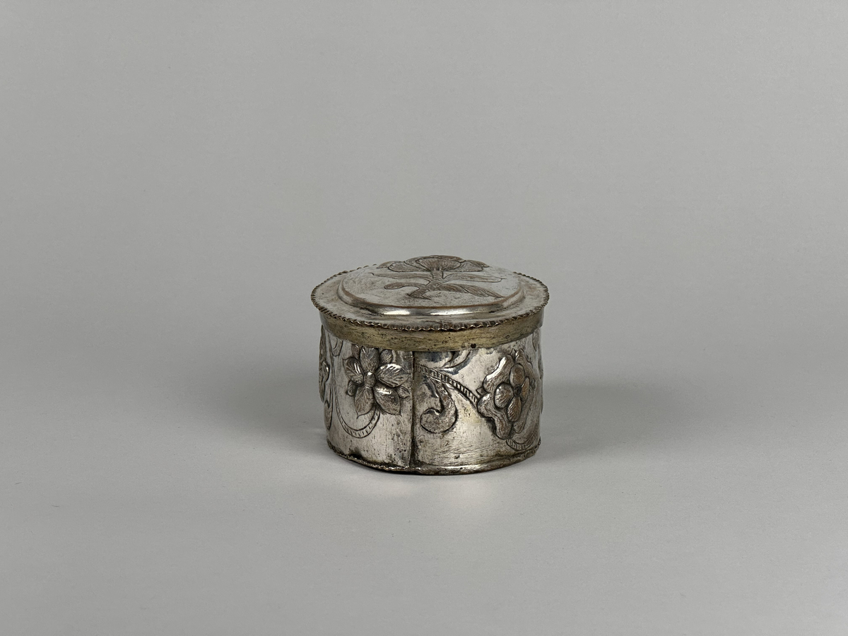 Oblatask tillverkad i försilvrad koppar, oval form med driven ornering. Lock dekorerad med en ros och sidorna med en girlang av rosor.