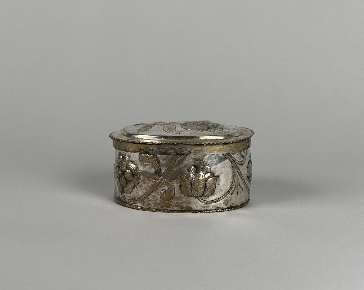 Oblatask tillverkad i försilvrad koppar, oval form med driven ornering. Lock dekorerad med en ros och sidorna med en girlang av rosor.