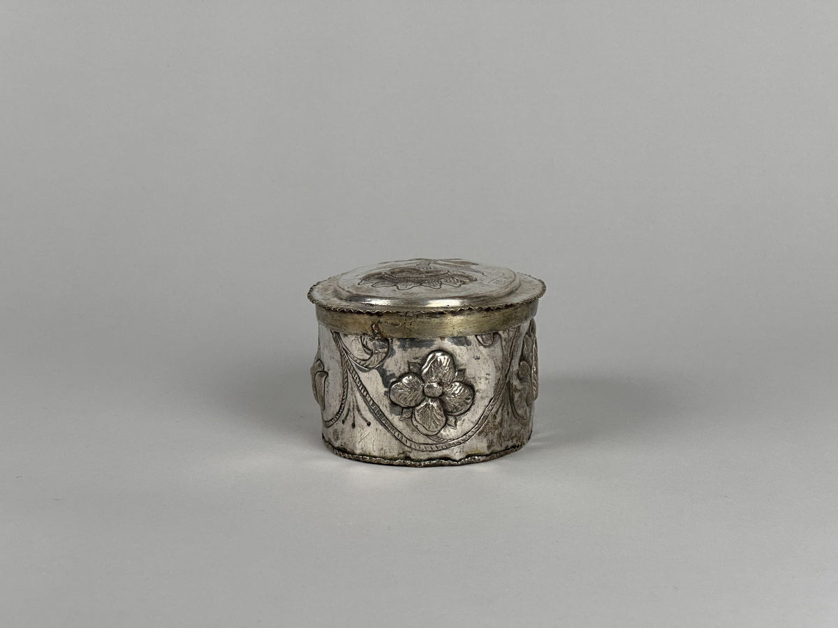 Oblatask tillverkad i försilvrad koppar, oval form med driven ornering. Lock dekorerad med en ros och sidorna med en girlang av rosor.