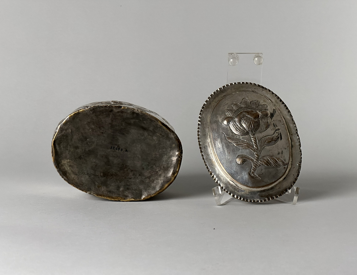 Oblatask tillverkad i försilvrad koppar, oval form med driven ornering. Lock dekorerad med en ros och sidorna med en girlang av rosor.