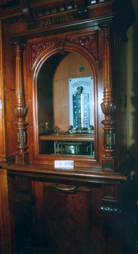 Vertikalstående, stor spilledåse av typen Polyphon, modell 54. Prod. nr. 3454. Maskinen bruker 63 cms plater.

Høyt skap i polert nøttetre med glassdør øverst og skuff nederst. Bak glassdøren ligger maskineriet og platearmen. Ved maskineriet ligger en lapp med bruksanvisning på tysk, engelsk og fransk. På skapetdøren, på hver side av glassplaten, står to rikt utskjærte dørsøyler. Øverst i hjørnene er det gjennombrudt akantus-utskjæringer. På høyre side av skapet er et myntinnkast som tar 5-øringer. Skuffen har to rikt utskjærte stolper og ornamentert metallhåndtak. Den åpnes på skrå og har plass til en rekke plater. Øverst i høyre hjørnet inne i skuffen er en mindre pengeskuff.

Til spilleren hører to små nøkler.