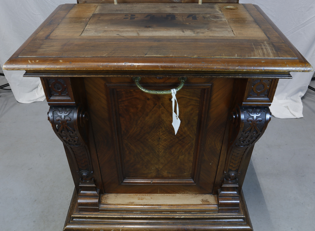 Vertikalstående, stor spilledåse av typen Polyphon, modell 54. Prod. nr. 3454. Maskinen bruker 63 cms plater.

Høyt skap i polert nøttetre med glassdør øverst og skuff nederst. Bak glassdøren ligger maskineriet og platearmen. Ved maskineriet ligger en lapp med bruksanvisning på tysk, engelsk og fransk. På skapetdøren, på hver side av glassplaten, står to rikt utskjærte dørsøyler. Øverst i hjørnene er det gjennombrudt akantus-utskjæringer. På høyre side av skapet er et myntinnkast som tar 5-øringer. Skuffen har to rikt utskjærte stolper og ornamentert metallhåndtak. Den åpnes på skrå og har plass til en rekke plater. Øverst i høyre hjørnet inne i skuffen er en mindre pengeskuff.

Til spilleren hører to små nøkler.