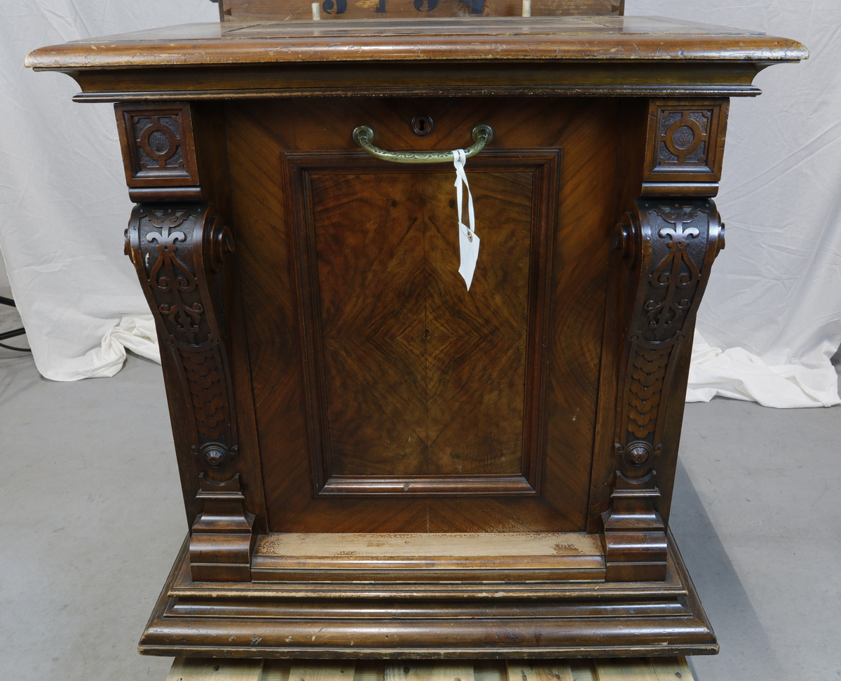 Vertikalstående, stor spilledåse av typen Polyphon, modell 54. Prod. nr. 3454. Maskinen bruker 63 cms plater.

Høyt skap i polert nøttetre med glassdør øverst og skuff nederst. Bak glassdøren ligger maskineriet og platearmen. Ved maskineriet ligger en lapp med bruksanvisning på tysk, engelsk og fransk. På skapetdøren, på hver side av glassplaten, står to rikt utskjærte dørsøyler. Øverst i hjørnene er det gjennombrudt akantus-utskjæringer. På høyre side av skapet er et myntinnkast som tar 5-øringer. Skuffen har to rikt utskjærte stolper og ornamentert metallhåndtak. Den åpnes på skrå og har plass til en rekke plater. Øverst i høyre hjørnet inne i skuffen er en mindre pengeskuff.

Til spilleren hører to små nøkler.