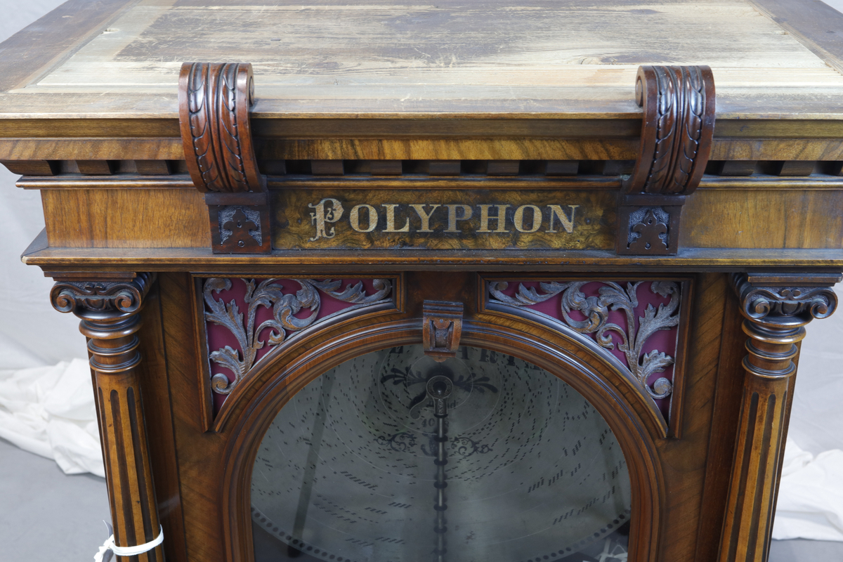 Vertikalstående, stor spilledåse av typen Polyphon, modell 54. Prod. nr. 3454. Maskinen bruker 63 cms plater.

Høyt skap i polert nøttetre med glassdør øverst og skuff nederst. Bak glassdøren ligger maskineriet og platearmen. Ved maskineriet ligger en lapp med bruksanvisning på tysk, engelsk og fransk. På skapetdøren, på hver side av glassplaten, står to rikt utskjærte dørsøyler. Øverst i hjørnene er det gjennombrudt akantus-utskjæringer. På høyre side av skapet er et myntinnkast som tar 5-øringer. Skuffen har to rikt utskjærte stolper og ornamentert metallhåndtak. Den åpnes på skrå og har plass til en rekke plater. Øverst i høyre hjørnet inne i skuffen er en mindre pengeskuff.

Til spilleren hører to små nøkler.