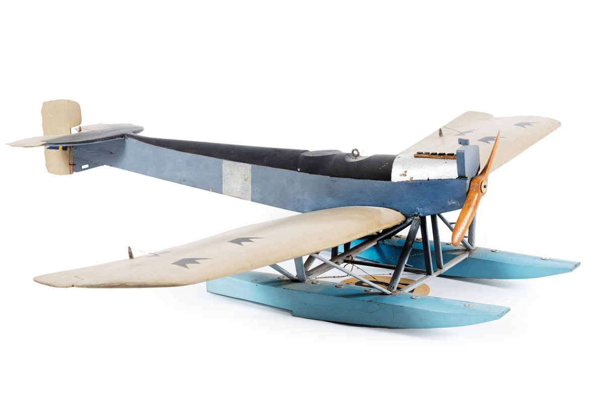 Modell av flygplan, Hansa-Brandenburg. Enmotorigt monoplan, två flottörer. Inbyggd flygkropp, placerad ovan vingarna. Breda och långa vingar med horisontala roder, vars ytterkant skjuta utanför vingarna. Stjärt med halvcirkelformad bäryta, två horisontala roder av oregelbunden form och med vågig akterkant, ett vertikalt roder. Stöttor och stag av metall. Gråmålat. Flygkropp svart- och blåmålad, flottörer blå. Motorhuv försilvrad.