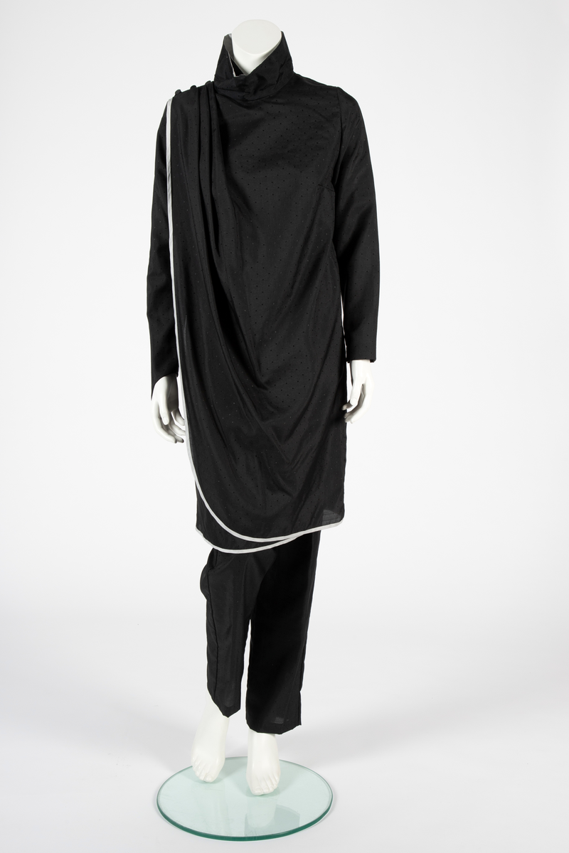 Ensemble i svart syntetisk silke, bestående av en lang jakke og en smal bukse med detaljer i lysegrå silke. Det matte svarte stoffet er tett besatt med ivevde, små, svarte blanke pletter. Det grå stoffet er umønstret. Jakken har et rett snitt med søm midt i ryggen og i hver side og når omtrent til knærne. Jakken har form som en omslagsjakke med under- og overstykke, begge i forsidens bredde, men venstre del er drapert fra høyre skulder, hvor det er knappet og er lett avrundet nederst. Kantet med en smal, grå bord. Oppstående halslinning, fôret med grått, knappet med trykknapper og hjørnet brettet ut. Isydde, lange smale ermer; høyre erme fortsetter i en spiss utover håndbaken og ender i en stropp som tres inn på langfingeren. Lang, smal bukse med sidesømmer. Livlining med én knapp foran og glidelås midt foran.
