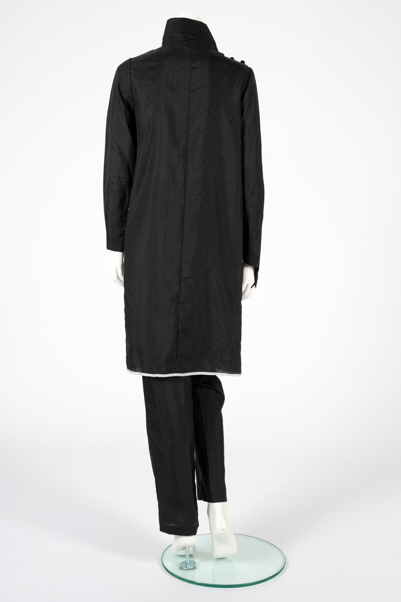 Ensemble i svart syntetisk silke, bestående av en lang jakke og en smal bukse med detaljer i lysegrå silke. Det matte svarte stoffet er tett besatt med ivevde, små, svarte blanke pletter. Det grå stoffet er umønstret. Jakken har et rett snitt med søm midt i ryggen og i hver side og når omtrent til knærne. Jakken har form som en omslagsjakke med under- og overstykke, begge i forsidens bredde, men venstre del er drapert fra høyre skulder, hvor det er knappet og er lett avrundet nederst. Kantet med en smal, grå bord. Oppstående halslinning, fôret med grått, knappet med trykknapper og hjørnet brettet ut. Isydde, lange smale ermer; høyre erme fortsetter i en spiss utover håndbaken og ender i en stropp som tres inn på langfingeren. Lang, smal bukse med sidesømmer. Livlining med én knapp foran og glidelås midt foran.