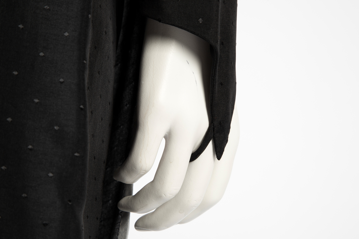 Ensemble i svart syntetisk silke, bestående av en lang jakke og en smal bukse med detaljer i lysegrå silke. Det matte svarte stoffet er tett besatt med ivevde, små, svarte blanke pletter. Det grå stoffet er umønstret. Jakken har et rett snitt med søm midt i ryggen og i hver side og når omtrent til knærne. Jakken har form som en omslagsjakke med under- og overstykke, begge i forsidens bredde, men venstre del er drapert fra høyre skulder, hvor det er knappet og er lett avrundet nederst. Kantet med en smal, grå bord. Oppstående halslinning, fôret med grått, knappet med trykknapper og hjørnet brettet ut. Isydde, lange smale ermer; høyre erme fortsetter i en spiss utover håndbaken og ender i en stropp som tres inn på langfingeren. Lang, smal bukse med sidesømmer. Livlining med én knapp foran og glidelås midt foran.