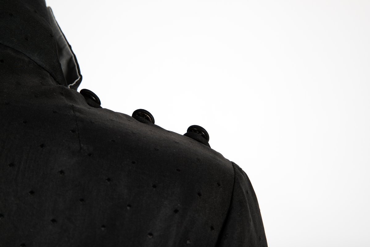 Ensemble i svart syntetisk silke, bestående av en lang jakke og en smal bukse med detaljer i lysegrå silke. Det matte svarte stoffet er tett besatt med ivevde, små, svarte blanke pletter. Det grå stoffet er umønstret. Jakken har et rett snitt med søm midt i ryggen og i hver side og når omtrent til knærne. Jakken har form som en omslagsjakke med under- og overstykke, begge i forsidens bredde, men venstre del er drapert fra høyre skulder, hvor det er knappet og er lett avrundet nederst. Kantet med en smal, grå bord. Oppstående halslinning, fôret med grått, knappet med trykknapper og hjørnet brettet ut. Isydde, lange smale ermer; høyre erme fortsetter i en spiss utover håndbaken og ender i en stropp som tres inn på langfingeren. Lang, smal bukse med sidesømmer. Livlining med én knapp foran og glidelås midt foran.