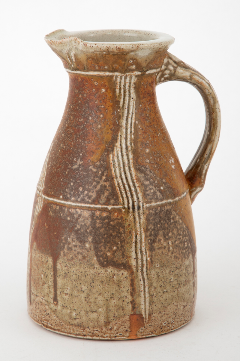 Mugge i steingods. Muggen har en sirkelrund bunn, omfangsrik nederst før den smalner inn mot munningen. Helt øverst vides den litt ut. Liten tut og kraftig hank. Det går vertikale linjer nedover korpus, risset inn i godset. Glasert i brunt med grønne, beige og grålige nyanser. Klar glasur innvendig.