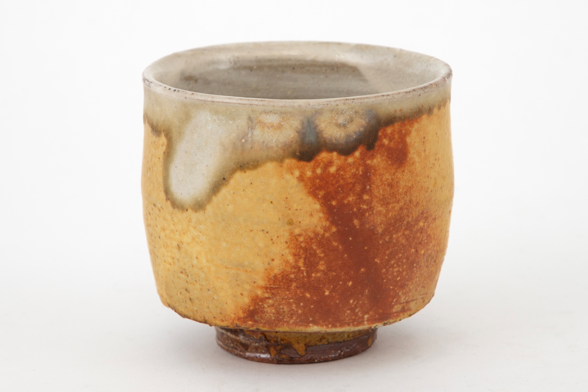 Kopp i steingods, sylinderform med liten fotring. Glasert i brunt med grønne, beige og grålige nyanser. Klar glasur innvendig.