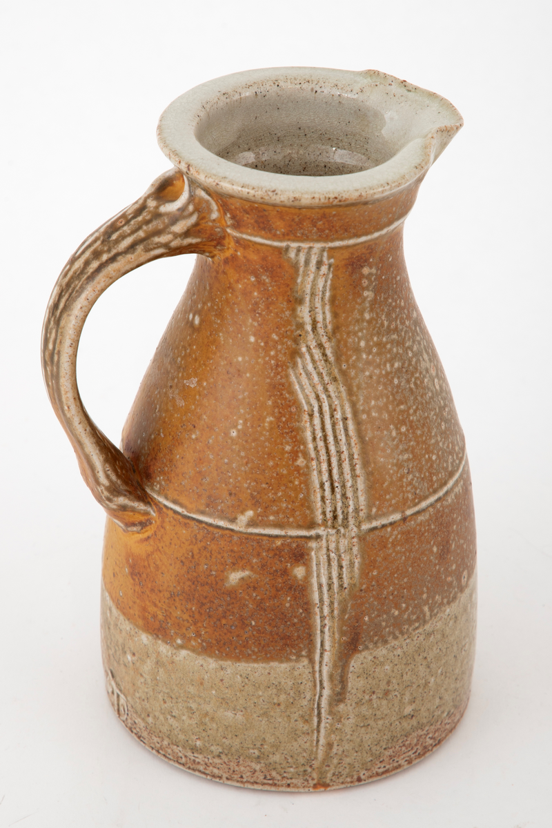 Mugge i steingods. Muggen har en sirkelrund bunn, omfangsrik nederst før den smalner inn mot munningen. Helt øverst vides den litt ut. Liten tut og kraftig hank. Det går vertikale linjer nedover korpus, risset inn i godset. Glasert i brunt med grønne, beige og grålige nyanser. Klar glasur innvendig.