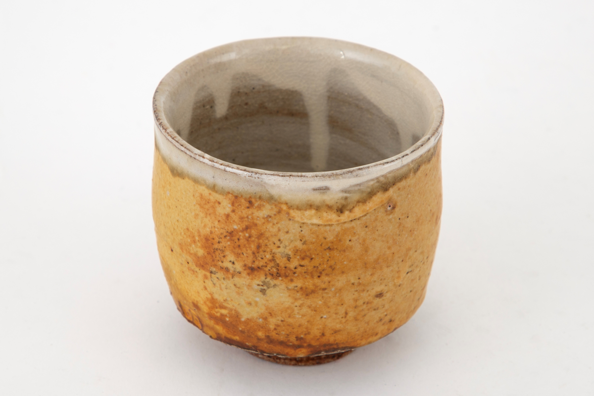 Kopp i steingods, sylinderform med liten fotring. Glasert i brunt med grønne, beige og grålige nyanser. Klar glasur innvendig.
