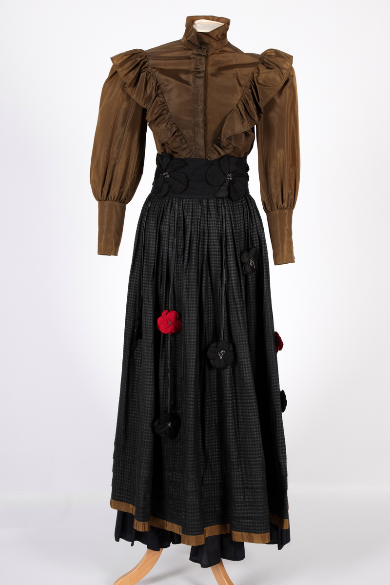 Smoking-/ gallaensemble bestående av en todelt kjole med belte, belteanheng, underskjørt og hårblomst. Bluse i syntetisk materiale som opprinnelig var svart, men over tid har blitt brunt. Blusen har vide, posete ermer og en bred rysjekant som løper over skuldrene og går ned i en spiss foran og bak. Oppstående halslinning med rysjekant. Skjult knappestolpe foran. Lange ermemansjetter med stoffkledde knapper. Langt skjørt med stor vidde i smårutete svart og grått bomullsstoff med kant av samme materiale som blusen nederst. Bredt, svart knyttebelte med stoffblomster dekorert med sølvfargede perler. Smalt rulourbelte i sateng med nedhengende blomster. Enkelte røde blomster er påsydd sekundært til bryllup. Underskjørt i svart, syntetisk materiale med kappe nederst.