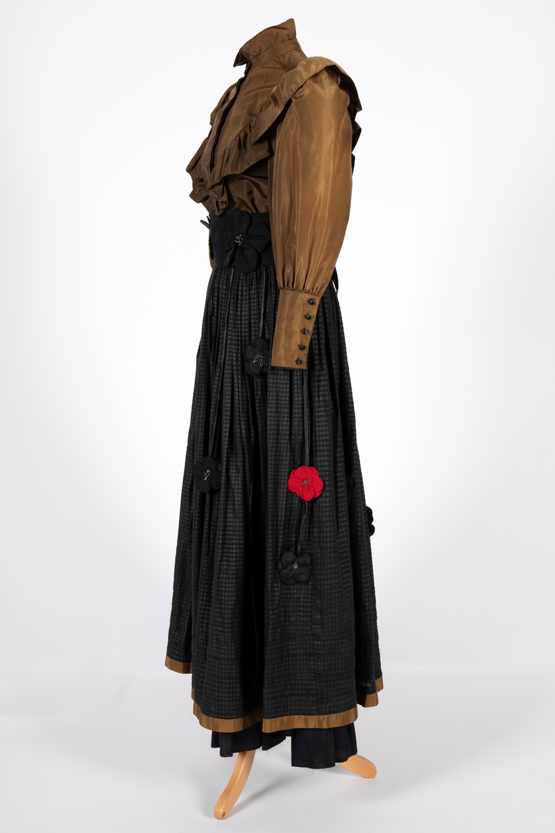 Smoking-/ gallaensemble bestående av en todelt kjole med belte, belteanheng, underskjørt og hårblomst. Bluse i syntetisk materiale som opprinnelig var svart, men over tid har blitt brunt. Blusen har vide, posete ermer og en bred rysjekant som løper over skuldrene og går ned i en spiss foran og bak. Oppstående halslinning med rysjekant. Skjult knappestolpe foran. Lange ermemansjetter med stoffkledde knapper. Langt skjørt med stor vidde i smårutete svart og grått bomullsstoff med kant av samme materiale som blusen nederst. Bredt, svart knyttebelte med stoffblomster dekorert med sølvfargede perler. Smalt rulourbelte i sateng med nedhengende blomster. Enkelte røde blomster er påsydd sekundært til bryllup. Underskjørt i svart, syntetisk materiale med kappe nederst.