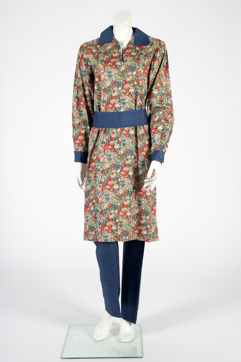 Buksedrakt bestående av tunikaoverdel, bukse og belte. Knelang tunikaoverdel i blomstret bomull med Liberty design og krage og mansjetter i blå silke med dekorative stikninger. Mansjettene kneppes med én knapp. Bukse i blå silke med smale bukseben. Knyttebelte i blå silke med stikninger.