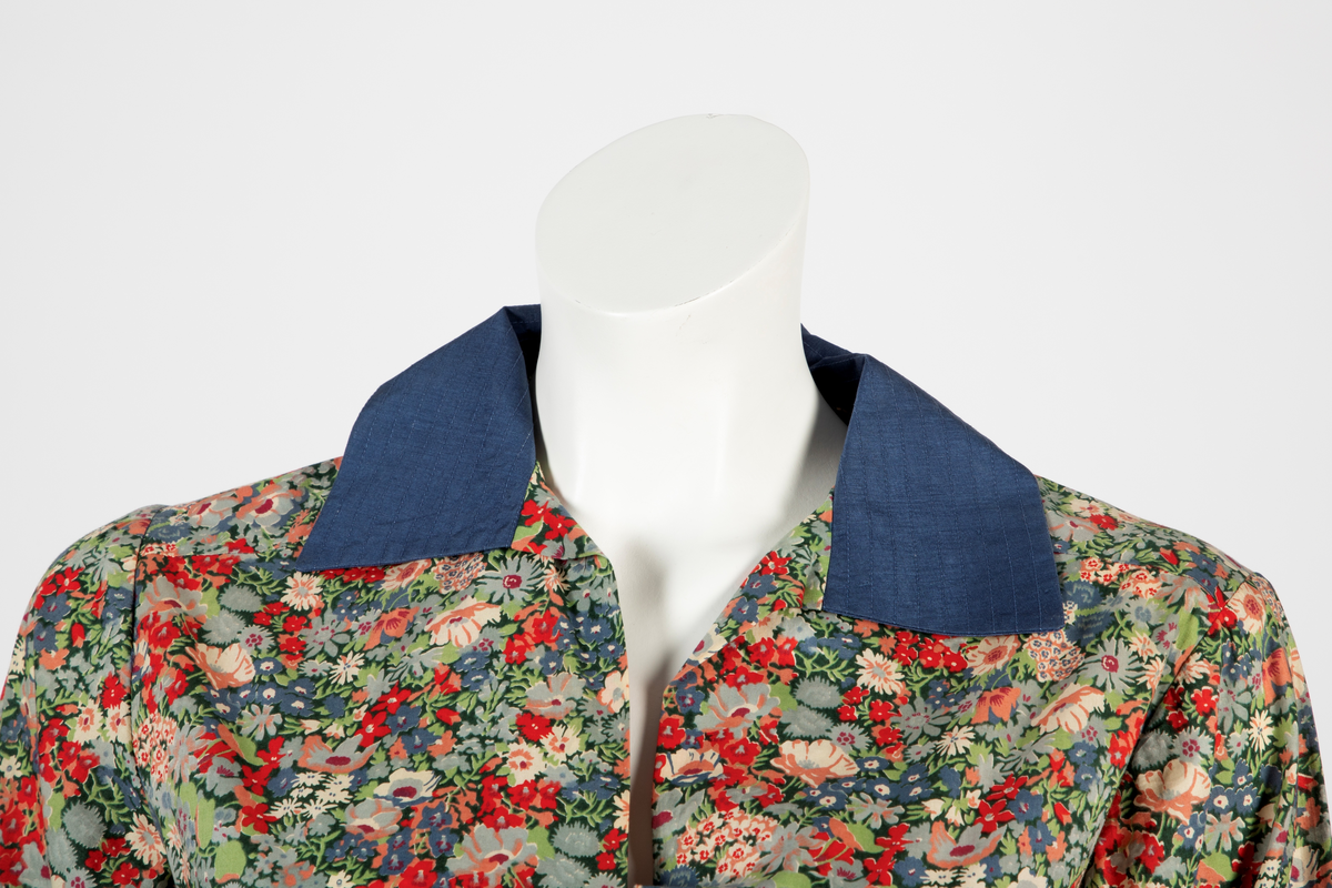 Buksedrakt bestående av tunikaoverdel, bukse og belte. Knelang tunikaoverdel i blomstret bomull med Liberty design og krage og mansjetter i blå silke med dekorative stikninger. Mansjettene kneppes med én knapp. Bukse i blå silke med smale bukseben. Knyttebelte i blå silke med stikninger.