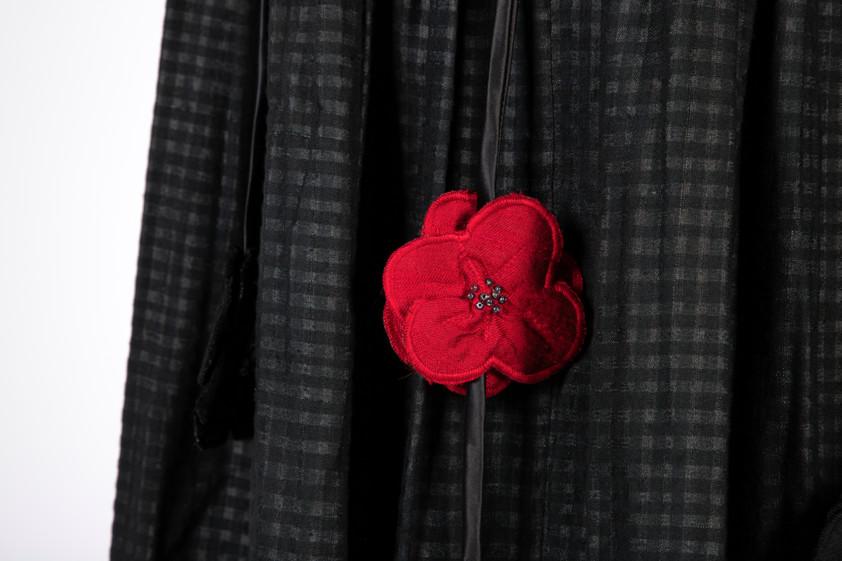 Smoking-/ gallaensemble bestående av en todelt kjole med belte, belteanheng, underskjørt og hårblomst. Bluse i syntetisk materiale som opprinnelig var svart, men over tid har blitt brunt. Blusen har vide, posete ermer og en bred rysjekant som løper over skuldrene og går ned i en spiss foran og bak. Oppstående halslinning med rysjekant. Skjult knappestolpe foran. Lange ermemansjetter med stoffkledde knapper. Langt skjørt med stor vidde i smårutete svart og grått bomullsstoff med kant av samme materiale som blusen nederst. Bredt, svart knyttebelte med stoffblomster dekorert med sølvfargede perler. Smalt rulourbelte i sateng med nedhengende blomster. Enkelte røde blomster er påsydd sekundært til bryllup. Underskjørt i svart, syntetisk materiale med kappe nederst.