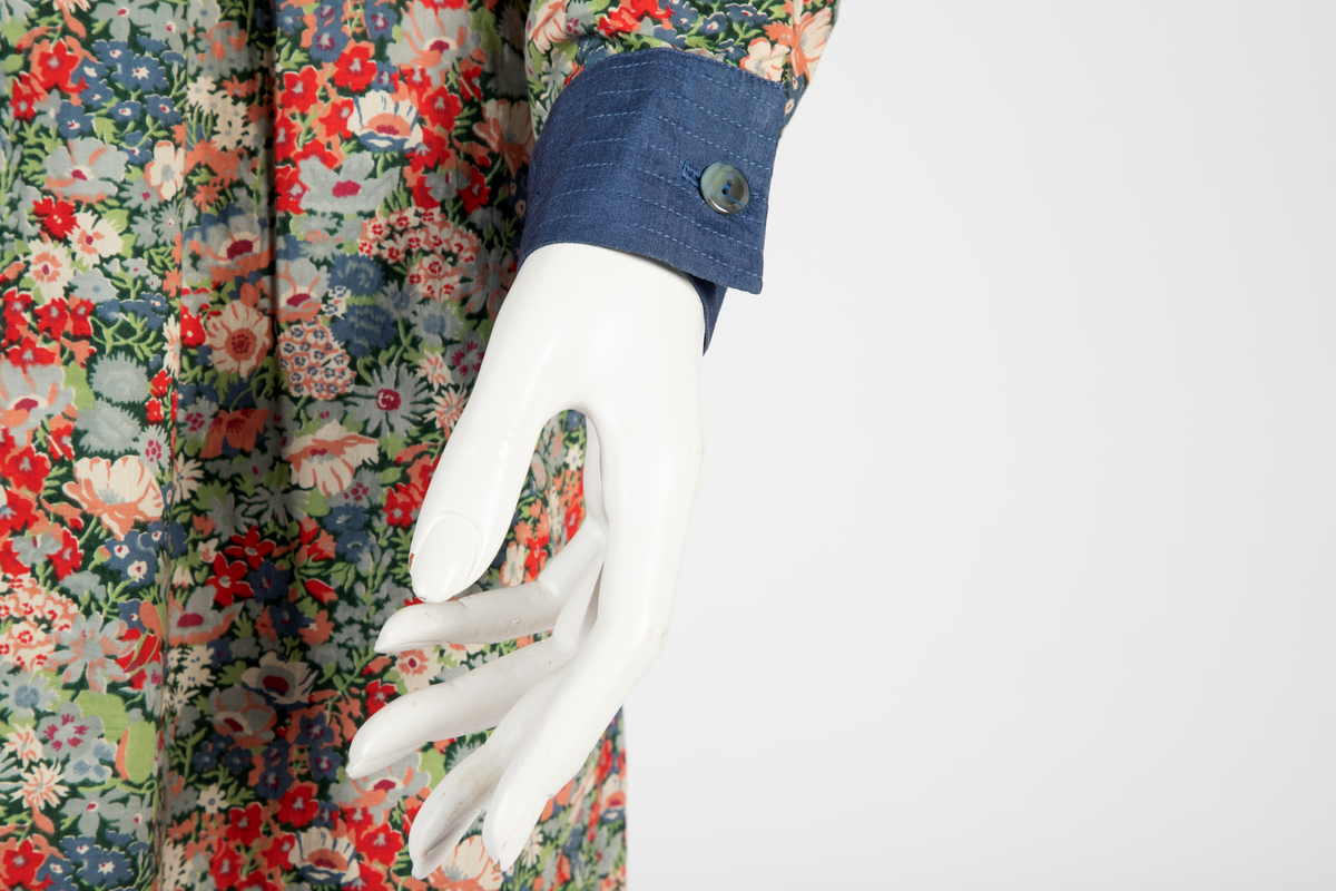 Buksedrakt bestående av tunikaoverdel, bukse og belte. Knelang tunikaoverdel i blomstret bomull med Liberty design og krage og mansjetter i blå silke med dekorative stikninger. Mansjettene kneppes med én knapp. Bukse i blå silke med smale bukseben. Knyttebelte i blå silke med stikninger.