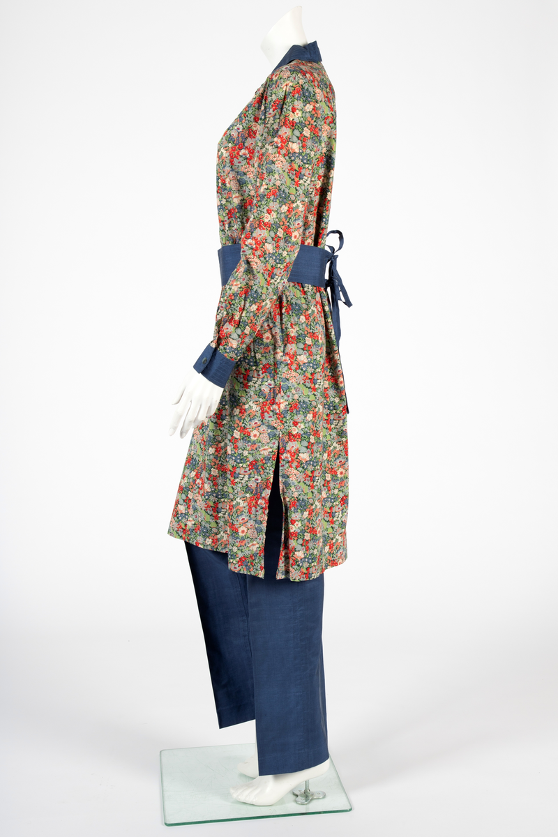 Buksedrakt bestående av tunikaoverdel, bukse og belte. Knelang tunikaoverdel i blomstret bomull med Liberty design og krage og mansjetter i blå silke med dekorative stikninger. Mansjettene kneppes med én knapp. Bukse i blå silke med smale bukseben. Knyttebelte i blå silke med stikninger.