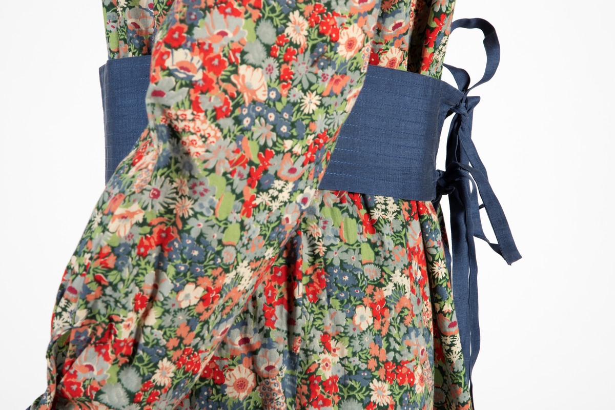 Buksedrakt bestående av tunikaoverdel, bukse og belte. Knelang tunikaoverdel i blomstret bomull med Liberty design og krage og mansjetter i blå silke med dekorative stikninger. Mansjettene kneppes med én knapp. Bukse i blå silke med smale bukseben. Knyttebelte i blå silke med stikninger.