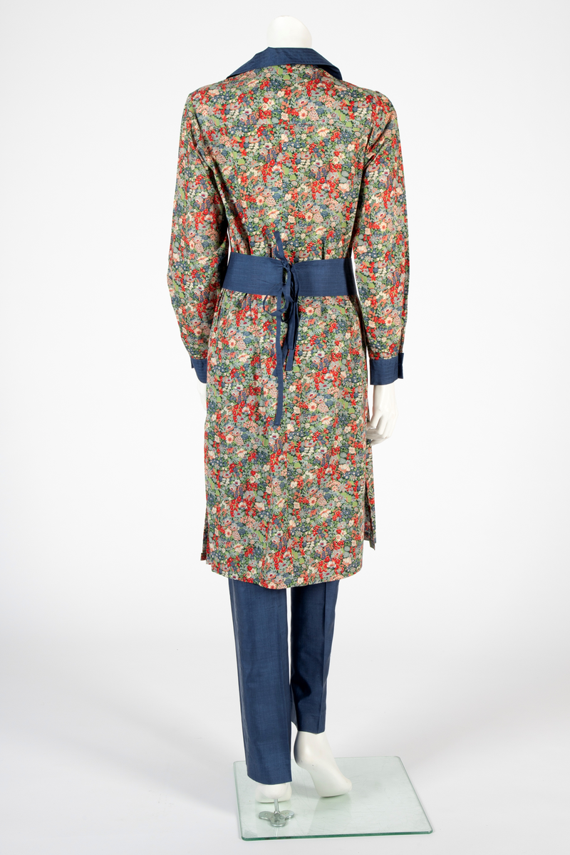 Buksedrakt bestående av tunikaoverdel, bukse og belte. Knelang tunikaoverdel i blomstret bomull med Liberty design og krage og mansjetter i blå silke med dekorative stikninger. Mansjettene kneppes med én knapp. Bukse i blå silke med smale bukseben. Knyttebelte i blå silke med stikninger.