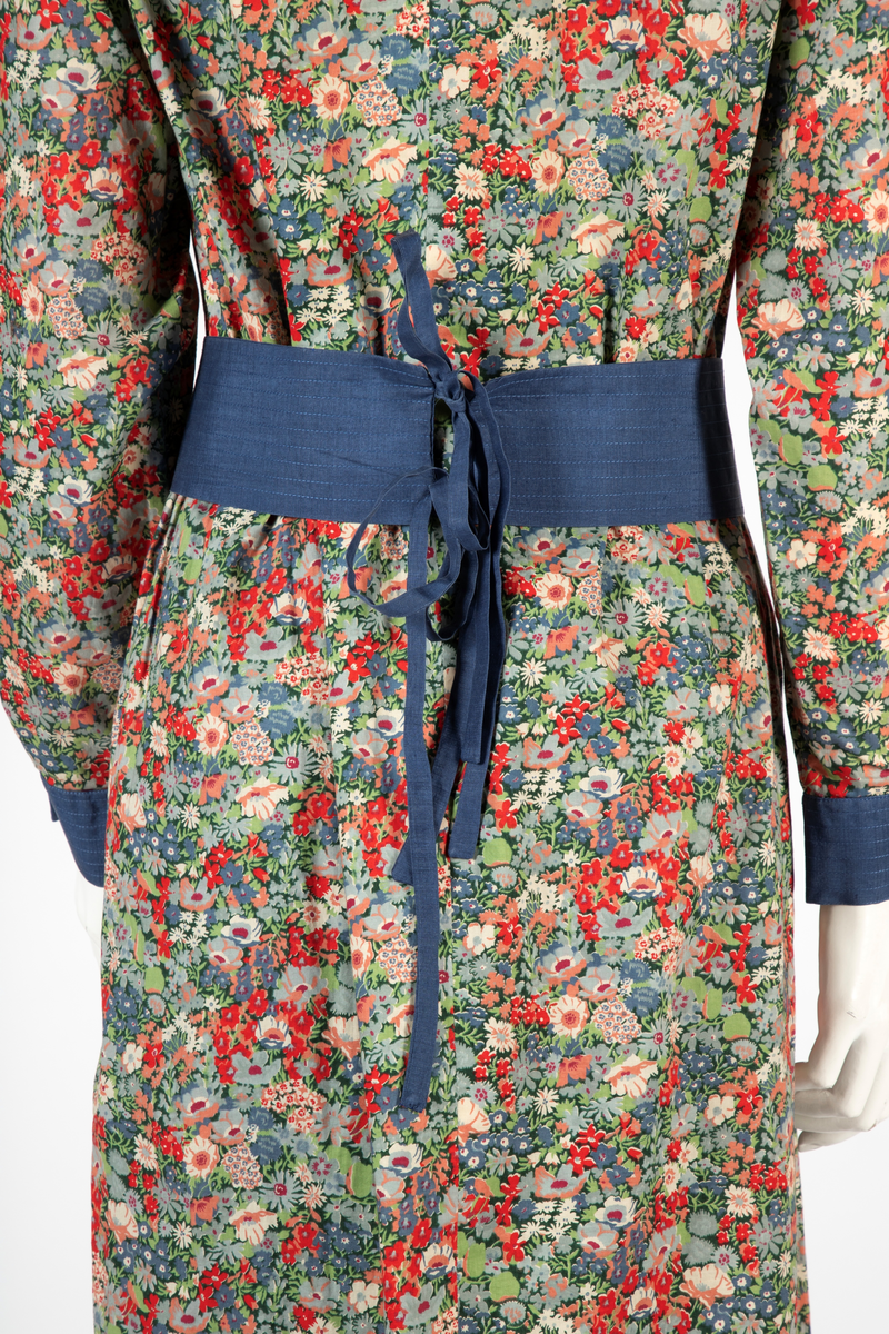 Buksedrakt bestående av tunikaoverdel, bukse og belte. Knelang tunikaoverdel i blomstret bomull med Liberty design og krage og mansjetter i blå silke med dekorative stikninger. Mansjettene kneppes med én knapp. Bukse i blå silke med smale bukseben. Knyttebelte i blå silke med stikninger.