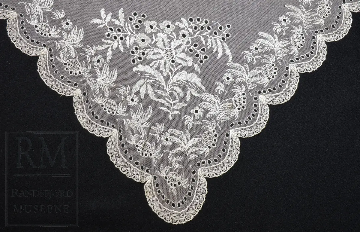 Kvadratisk form, med tungekant. Kantet med kniplingblonde. Tyllbroderi: blomsterranke langs sidene. Større hjørnemotiv i fire hjørner, der et er vesentlig større.  