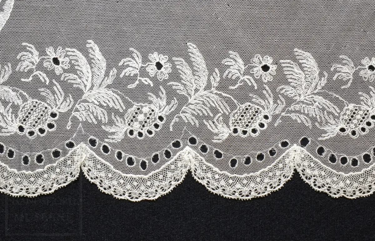 Kvadratisk form, med tungekant. Kantet med kniplingblonde. Tyllbroderi: blomsterranke langs sidene. Større hjørnemotiv i fire hjørner, der et er vesentlig større.  