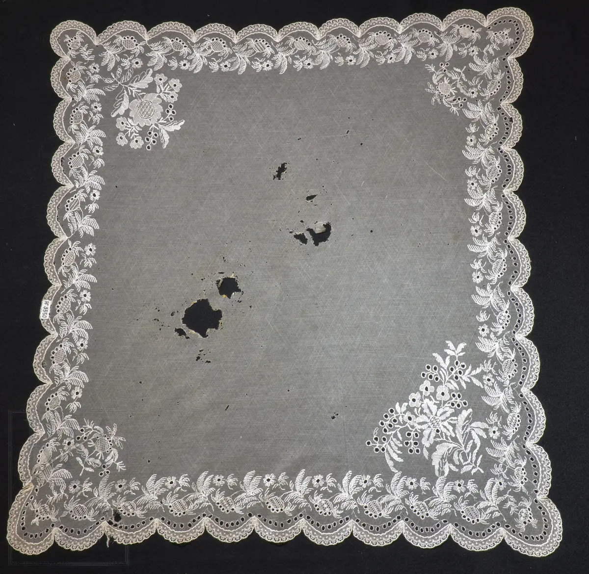 Kvadratisk form, med tungekant. Kantet med kniplingblonde. Tyllbroderi: blomsterranke langs sidene. Større hjørnemotiv i fire hjørner, der et er vesentlig større.  