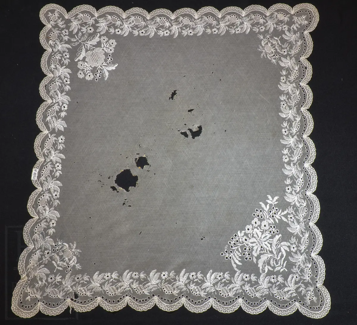 Kvadratisk form, med tungekant. Kantet med kniplingblonde. Tyllbroderi: blomsterranke langs sidene. Større hjørnemotiv i fire hjørner, der et er vesentlig større.  