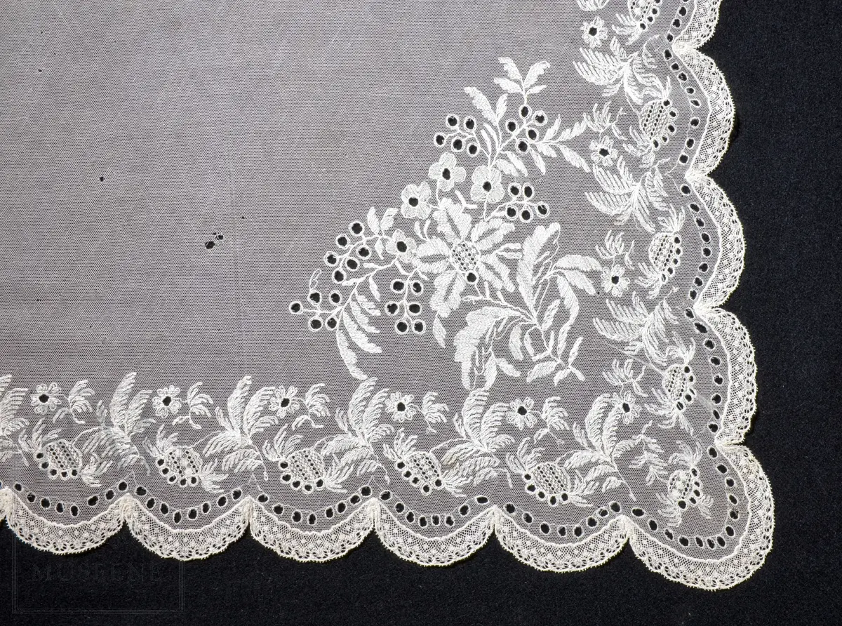 Kvadratisk form, med tungekant. Kantet med kniplingblonde. Tyllbroderi: blomsterranke langs sidene. Større hjørnemotiv i fire hjørner, der et er vesentlig større.  