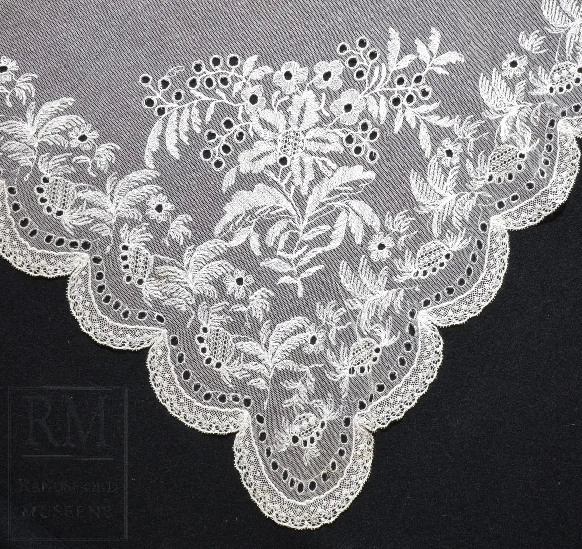 Kvadratisk form, med tungekant. Kantet med kniplingblonde. Tyllbroderi: blomsterranke langs sidene. Større hjørnemotiv i fire hjørner, der et er vesentlig større.  